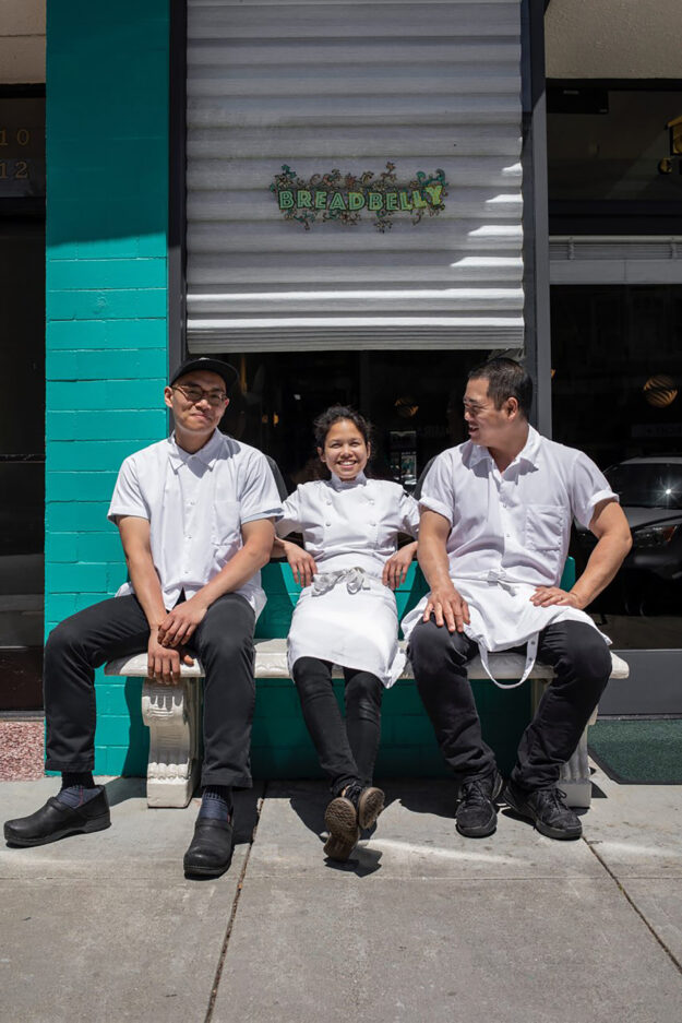 Breadbelly co-owners (left to right) Clement Hsu, Katherine Campecino-Wong, and James Wong sit in front of their storefront, a teal brick building with roll-down shutters with "Breadbelly" spelled out in green letters with flowery accents.